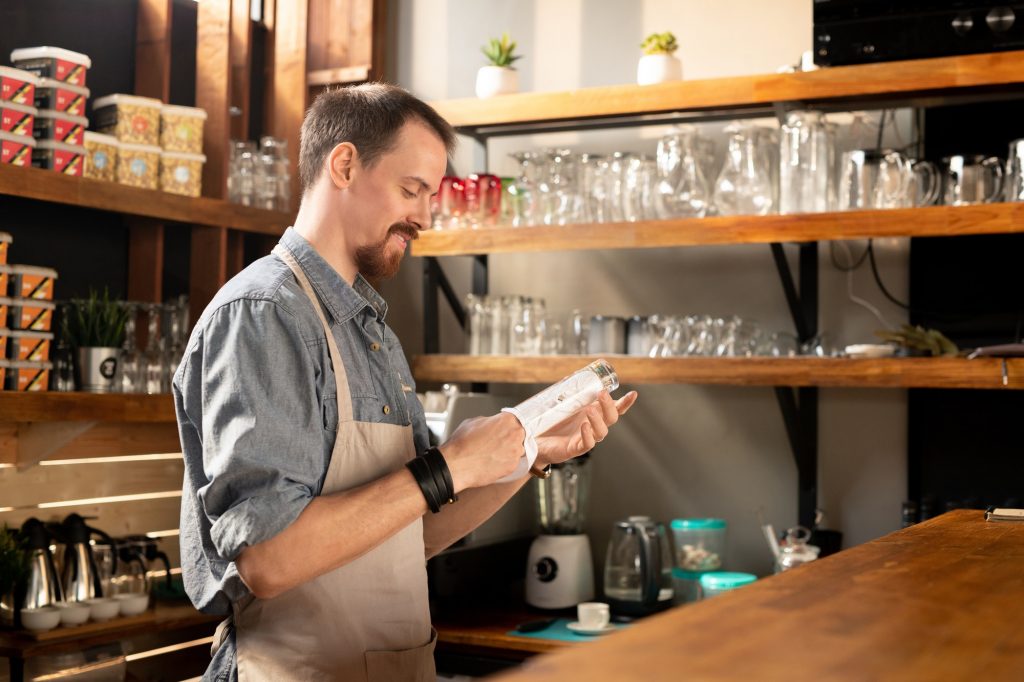 Happy young bearded waiter in apron wiping glass with napkin at Darren Yaw foo hoe